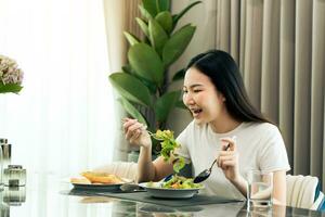 jeune femme asiatique souriante alors qu'elle prend une salade sur une assiette et mange joyeusement à la maison. photo