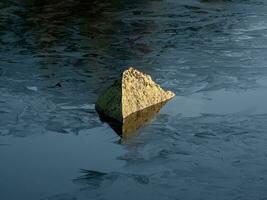 seul Roche dans congelé étang. une pierre cette est moitié submergé dans Matin congelé l'eau et exposé à le lumière du soleil. photo