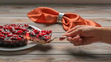 tarte avec baies, framboises, des fraises, groseilles, sur une blanc assiette, un femme main en portant une pièce de gâteau sur une spatule. sur une en bois Contexte photo