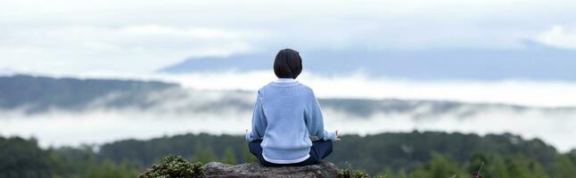 panorama retour vue de femme dans sweat à capuche est de façon relaxante pratiquant méditation yoga à le Haut de Montagne avec brouillard et brouillard dans été à atteindre bonheur de interne paix sagesse pour en bonne santé esprit et âme photo