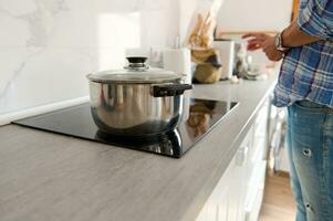 détails sur une inoxydable acier casserole sur noir verre surface de un électrique le fourneau dans une moderne blanc minimaliste cuisine photo