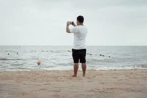 une relaxant homme permanent sur le plage et en utilisant une téléphone intelligent prend une photo sur le plage