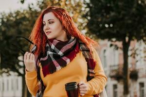 jeune femme rousse en pull jaune et lunettes de soleil tient une tasse thermo avec du café dans ses mains. dame se promène dans la ville et le parc le jour d'automne ensoleillé. photo