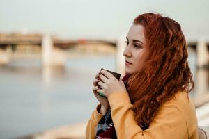 portrait de jeune femme rousse sur fond de rivière et de pont de la ville. elle boit du café dans une tasse en papier et profite de la vue d'automne. photo