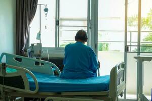 un personnes âgées asiatique Masculin patient est assis sur une hôpital lit attendre pour une médecin à vérifier sur lui pendant le sien hôpital rester. doux et sélectif se concentrer. photo