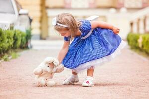 le peu fille sur le rue en jouant avec une jouet. bébé mettre jouet lapin sur le sol. une fille dans une luxuriant bleu robe en marchant dans le ville kyiv Ukraine. photo