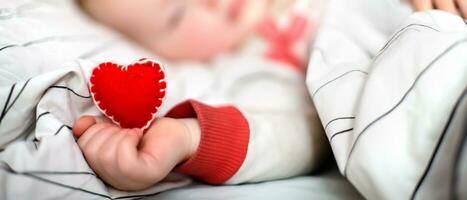 peu enfant dort dans lit. rouge cœur dans enfant main. la Saint-Valentin journée pour Parents avec les enfants. l'amour pour une enfant est concept. bébé a en bonne santé cœur. photo