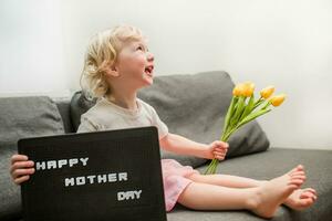 peu fille détient une bouquet de Jaune tulipes et une signe cette dit content de la mère journée. enfant donne fleurs à mère pour vacances. photo