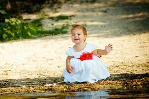 le peu fille dans une blanc robe séance sur le rivière banque. une enfant en jouant près le l'eau. le enfant sourit. enfant grimaça et fermé le sien yeux. photo