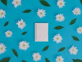 directement au dessus vue de Vide carnet avec blanc fleurs et vert feuilles sur bleu Contexte. photo