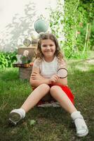 content souriant fille de sept ans vieux est assis sur herbe en plein air près école fournitures. globe, livres, des lettres et alarme l'horloge sur le Contexte de une blanc mur. photo