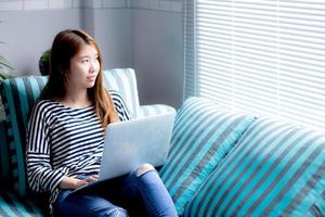 belle de portrait jeune femme asiatique utilisant un ordinateur portable pour les loisirs sur un canapé dans le salon, fille travaillant en ligne avec un ordinateur portable indépendant avec un concept d'entreprise de communication heureux.