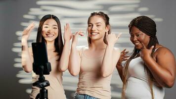 de bonne humeur femmes dansant et agissant idiot sur caméra, en utilisant téléphone intelligent dans studio et ayant amusement. diverse groupe de les filles profiter Danse et posant pour soi l'amour un d campagne, corps confiance. photo