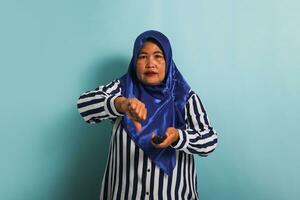 un malheureux âge moyen asiatique femme dans une bleu hijab et une rayé chemise est donnant une pouces vers le bas à une mal la télé montrer. elle est isolé sur une bleu Contexte photo