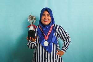 une content âge moyen asiatique femme d'affaires dans une bleu hijab et une rayé chemise est montrant un vide blanc médaille tandis que en portant une argent trophée, célébrer sa succès, isolé sur une bleu Contexte photo