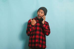 excité Jeune asiatique homme avec une bonnet chapeau et une rouge plaid flanelle chemise est montrer du doigt le sien doigt à le la gauche à copie espace avec pouce tandis que parlant sur le sien mobile téléphone, isolé sur une bleu Contexte photo