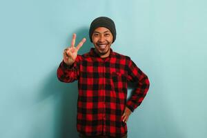 attrayant Jeune asiatique homme avec une bonnet chapeau et une rouge plaid flanelle chemise pose pour le caméra avec une sourire sur le sien visage et spectacles une paix geste tandis que permanent contre une bleu Contexte photo