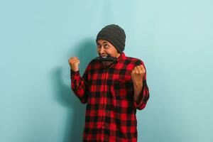 marrant Jeune asiatique homme avec une bonnet chapeau et rouge plaid flanelle chemise mordant une la télé télécommande, sentiment en colère tandis que en train de regarder une des sports Jeu sur la télé, isolé sur une bleu Contexte photo