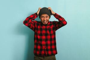 un en colère Jeune asiatique homme avec une bonnet chapeau et une rouge plaid flanelle chemise attrape le sien diriger, sentiment mécontent, bouleversé, et déçu. il est isolé sur une bleu Contexte photo