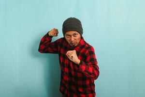 un en colère, furieux Jeune asiatique homme avec une bonnet chapeau et une rouge plaid flanelle chemise est prêt à lutte, avec une défensive poing geste, sentiment furieux et bouleversé. il est isolé sur une bleu Contexte photo