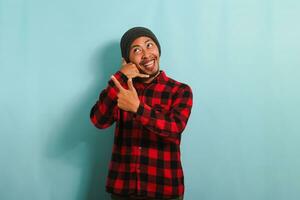 un excité Jeune asiatique homme portant une bonnet chapeau et une rouge plaid flanelle chemise fait du une appel moi geste, montrer du doigt à le la gauche espace de copie, isolé sur une bleu Contexte photo