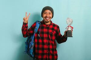 excité Jeune asiatique homme étudiant portant une sac à dos, bonnet chapeau, et rouge plaid flanelle chemise, fabrication une la victoire geste tandis que en portant une trophée et réjouissance dans réalisation, isolé sur une bleu Contexte photo