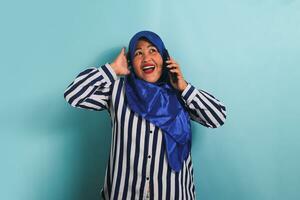 un excité âge moyen asiatique femme dans une bleu hijab et une rayé chemise est en riant tandis que parlant sur sa téléphone portable. elle est isolé sur une bleu Contexte. photo
