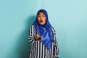 une sous le choc âge moyen asiatique femme dans une bleu hijab est en train de regarder télévision, montrer du doigt le la télé éloigné manette vers le caméra, et commutation canaux avec un ouvert bouche, isolé sur une bleu Contexte photo