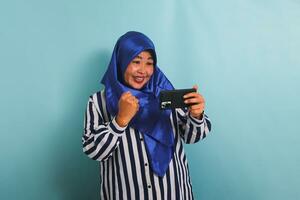 excité âge moyen asiatique femme dans une bleu hijab et une rayé chemise, en jouant une Jeu sur mobile téléphone, isolé sur une bleu Contexte. photo