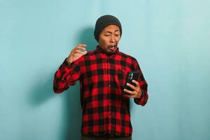 marrant Jeune asiatique homme avec une bonnet chapeau et une rouge plaid flanelle chemise est dans choc et surpris tandis que en train de lire mal nouvelles sur le sien téléphone intelligent, isolé sur une bleu Contexte. photo