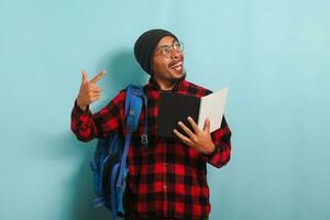 excité Jeune asiatique étudiant est en portant livres et montrer du doigt tandis que permanent contre une bleu Contexte photo