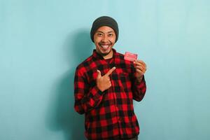 excité Jeune asiatique homme s montrer du doigt à le sien banque crédit cartes dans le sien mains isolé sur bleu Contexte photo