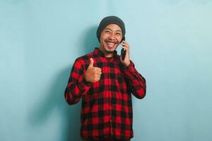 excité Jeune asiatique homme avec une bonnet chapeau et une rouge plaid flanelle chemise est montrant une les pouces en haut tandis que parlant sur le sien mobile téléphone, isolé sur une bleu Contexte photo