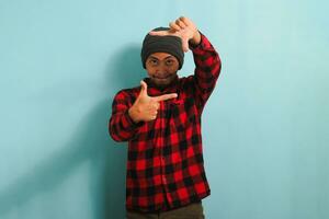 excité Jeune asiatique homme avec une bonnet chapeau et une rouge plaid flanelle chemise est fabrication une image Cadre geste, prétendre à concentrer comme une photographe, isolé sur une bleu Contexte photo