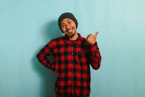 excité Jeune asiatique homme avec une bonnet chapeau et rouge plaid flanelle chemise est montrer du doigt de côté avec le sien pouce, indiquant le copie espace, isolé sur une bleu Contexte photo