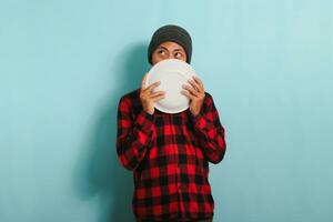 faim Jeune asiatique homme avec une bonnet chapeau et une rouge plaid flanelle chemise est à la recherche de côté, en pensant à propos quoi à manger, tandis que en portant un vide blanc assiette dans le sien main, isolé sur une bleu Contexte photo