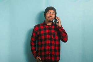 sur de soi Jeune asiatique homme avec une bonnet chapeau et une rouge plaid flanelle chemise est souriant à le caméra tandis que parlant sur le sien téléphone intelligent, isolé sur une bleu Contexte photo