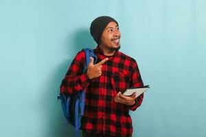 une souriant Jeune asiatique étudiant avec une bonnet chapeau et une rouge plaid flanelle chemise, portant une sac à dos, est montrer du doigt doigt à le droite à copie espace tandis que permanent contre une bleu Contexte photo