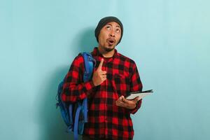 un excité souriant Jeune asiatique étudiant avec une bonnet chapeau et une rouge plaid flanelle chemise, portant une sac à dos, est montrer du doigt doigt en haut à copie espace tandis que permanent contre une bleu Contexte photo