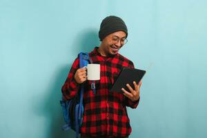 une Jeune asiatique Masculin étudiant avec une bonnet chapeau et une rouge plaid flanelle chemise, portant une sac à dos et lunettes, prend une café Pause après conférences tandis que permanent contre une bleu Contexte photo