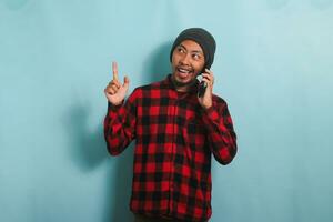 excité Jeune asiatique homme avec une bonnet chapeau et une rouge plaid flanelle chemise est montrer du doigt vers le haut à copie espace tandis que parlant sur le sien mobile téléphone, isolé sur une bleu Contexte photo