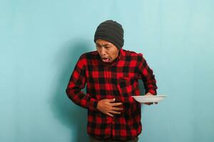 Jeune asiatique homme avec une bonnet chapeau et une rouge plaid flanelle chemise est sentiment nauséeux tandis que en portant blanc plaque, isolé sur une bleu Contexte photo