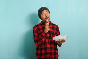 pensif Jeune asiatique homme avec bonnet chapeau et rouge plaid flanelle chemise en portant une cuillère et un vide blanc assiette dans le sien main, en pensant à propos quoi à manger, isolé sur une bleu Contexte photo