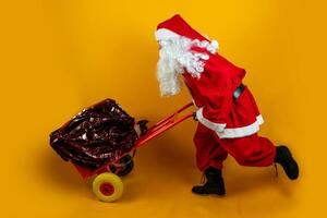 Père Noël claus est fonctionnement avec une sac plein de présente sur une métal chariot sur Orange Contexte photo