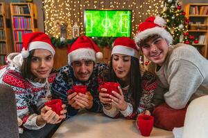 groupe de quatre Jeune copains grillage avec rouge tasses habillé dans rouge Chapeaux et Noël pulls molletonnés photo