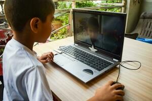 peu garçon séance à table en utilisant portable pour en ligne classe dans classe 1, enfant en train d'étudier sur portable de Accueil pour distance apprentissage en ligne éducation, école garçon les enfants mode de vie concept photo