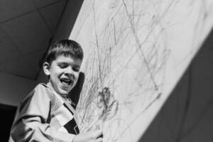 en riant enfant dessine sur le mur avec craie dans le sien chambre. le garçon est engagé dans la créativité à maison. noir et blanc photo