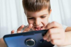 une de bonne humeur enfant dans une blanc T-shirt pièces Jeux sur le téléphone à maison. content garçon regards à le sien téléphone intelligent. photo
