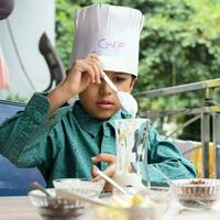 mignonne Indien chef garçon en train de préparer sundae plat comme une partie de non Feu cuisine lequel comprend vanille la glace crème, lutin, coco poudre, fraîchement haché des fruits et fraise sirop. peu enfant en train de préparer nourriture photo