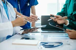 médical équipe ayant une réunion avec médecins dans blanc laboratoire manteaux et chirurgical gommages assise à une table discuter une les patients travail en ligne en utilisant des ordinateurs photo